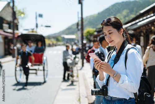 portrait of pretty Asian female visitor in japan is using mobile phone on the sidewalk at Arashiyama main street with running rickshaw in the blurred background