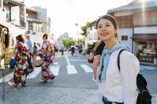 smiling asian girl is looking into distance on street with females wearing kimono at background while visiting historic district Kyoto, japan on autumn sunny day