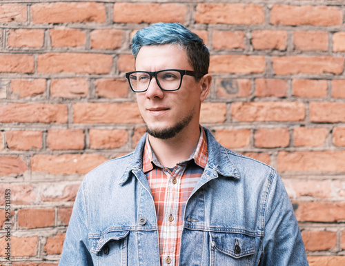 Stylish and fashionable handsome young man with blue hair wearing in jeans jacket standing on background of brick wall.