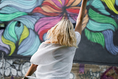 Pretty young woman with blond hair standing on a street with the graffiti wall background. View from the back