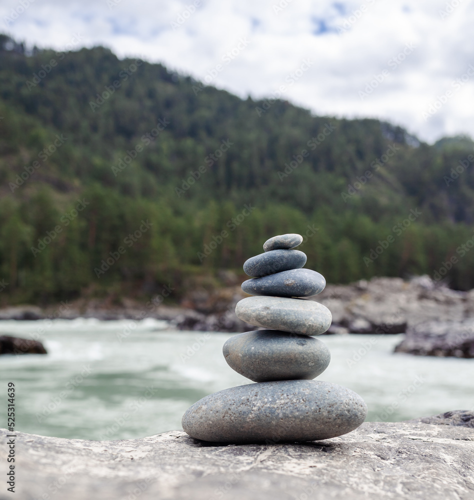A pyramid of bare stones stacked on top of each other. Stones stacked in the shape of a pyramid on the riverbank against the background of mountains as balance and balance in nature, Zen, Buddhism. A pyramid of bare stones stacked on top of each other. Stones stacked in the shape of a pyramid on the riverbank against the background of mountains as balance and balance in nature, Zen, Buddhism.