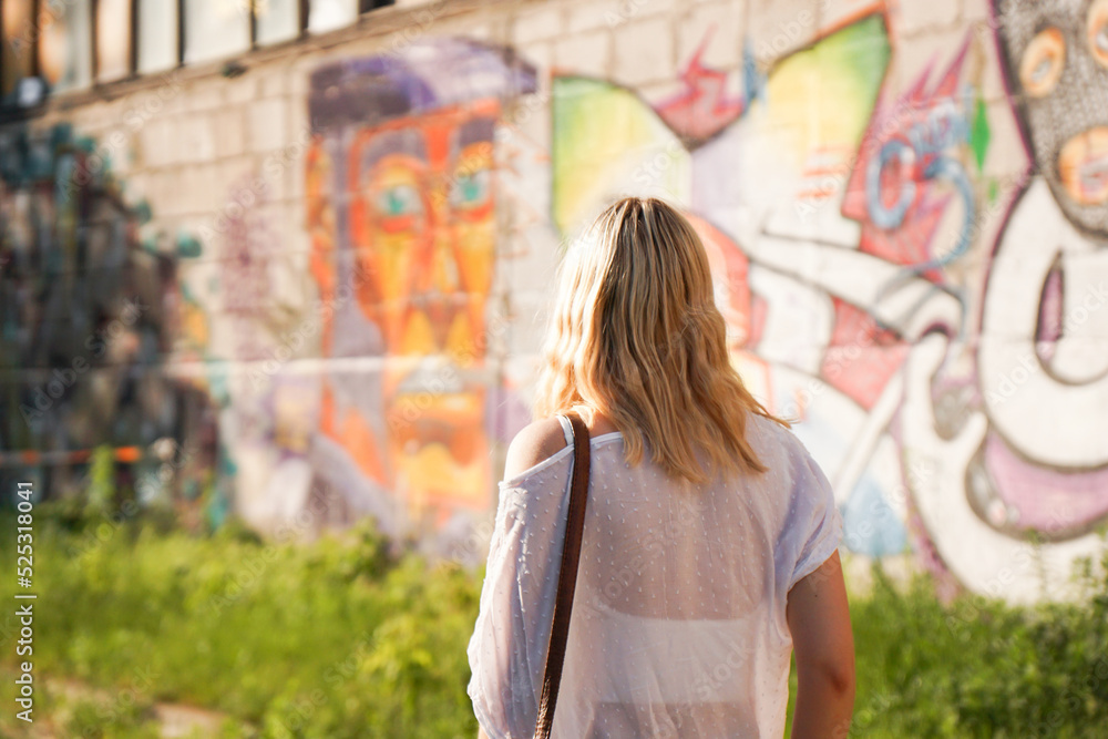 © brillianata - Pretty young woman with blond hair standing on a street with the graffiti wall background. View from the back
