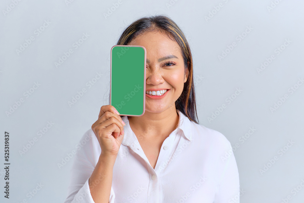Smiling young Asian woman covering half face with blank screen mobile phone, recommending mobile app isolated over white background Smiling young Asian woman covering half face with blank screen mobile phone, recommending mobile app isolated over white background