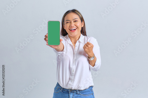 Excited young Asian woman showing blank screen mobile phone and making winner gesture isolated over white background