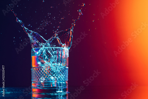 Whiskey glass with ice cubes and water splashes standing on mirror surface over gradient black orange background in neon light. Art, beauty, drinks