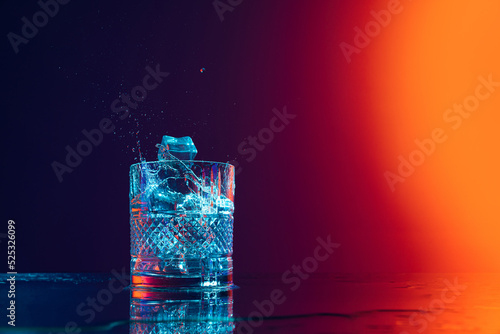 Ice cube falls into a transparent glass of water standing on mirror surface over gradient black orange background in neon light. Art, beauty, drinks