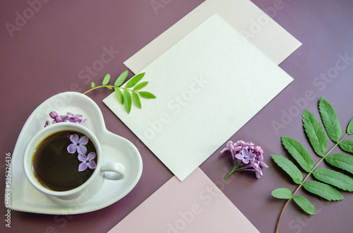 close-up diagonal Mockup of white cards and a pink envelope on a dark purple background with leaves and lilac flowers with a coffee cup and heart-shaped saucer