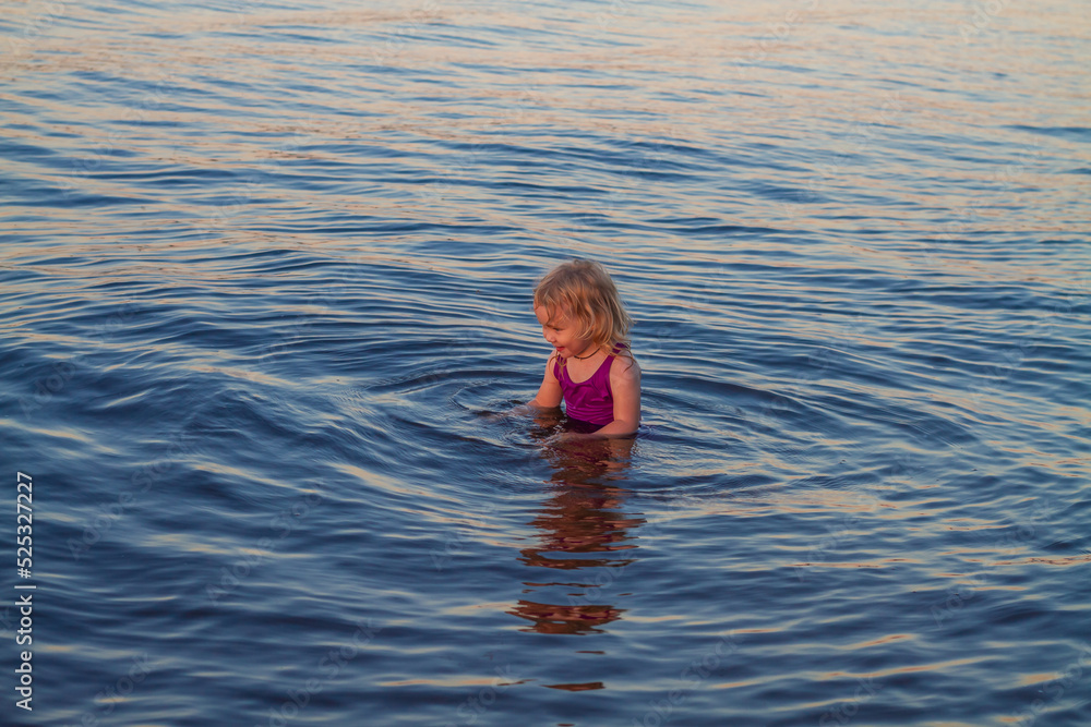 Gentle baby blonde girl 3 years old swims in the water in the river, lake, sea in summer in a purple swimsuit