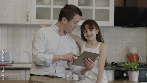 Happy father and his little daughter are using tablet computer at the dinner table in the kitchen, checking bank accounts, shopping online, checking social networks, ordering food from restaurant. photo