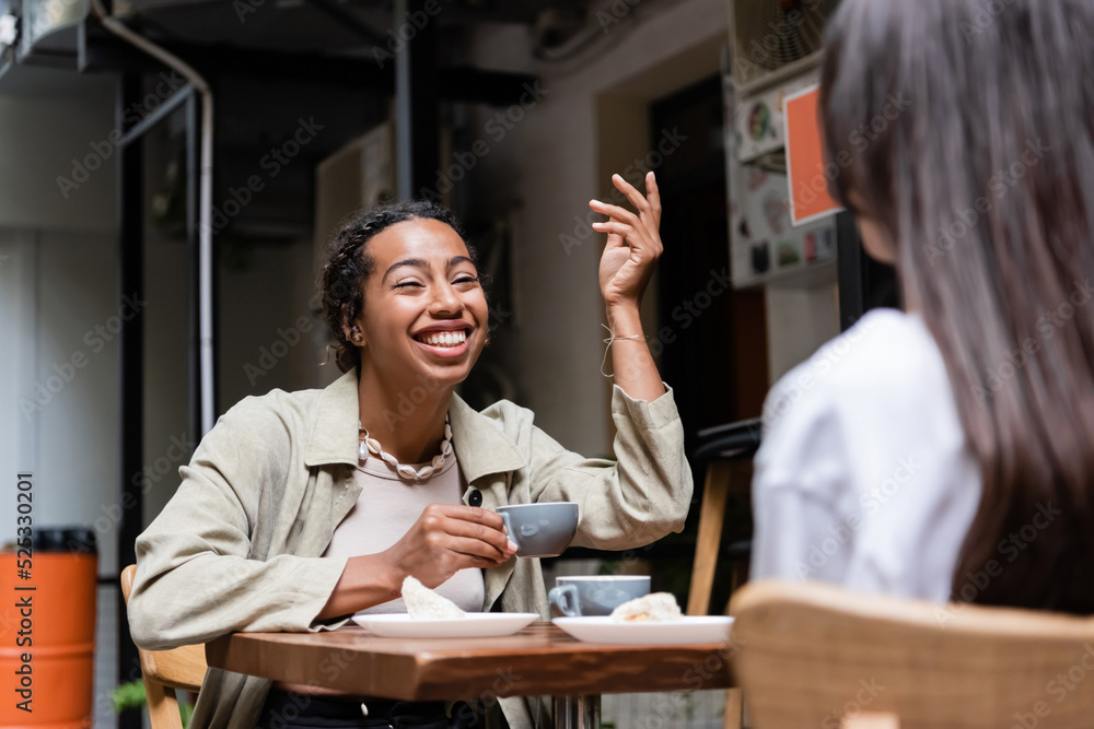 African american woman holding coffee cup and talking to blurred girlfriend on terrace of cafe African american woman holding coffee cup and talking to blurred girlfriend on terrace of cafe