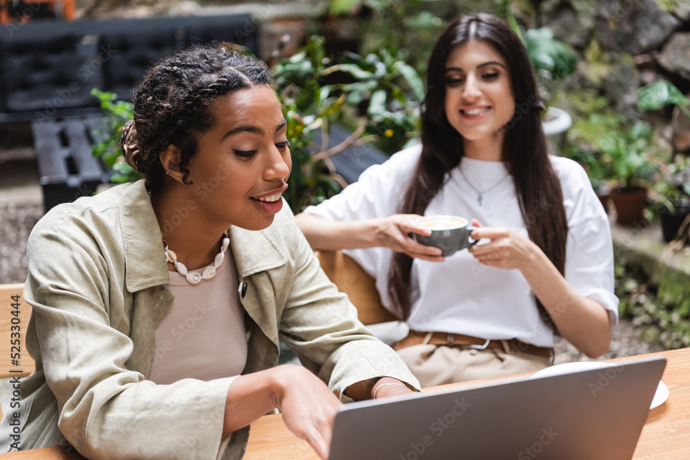 Smiling african american woman using laptop near blurred friend with coffee in outdoor cafe