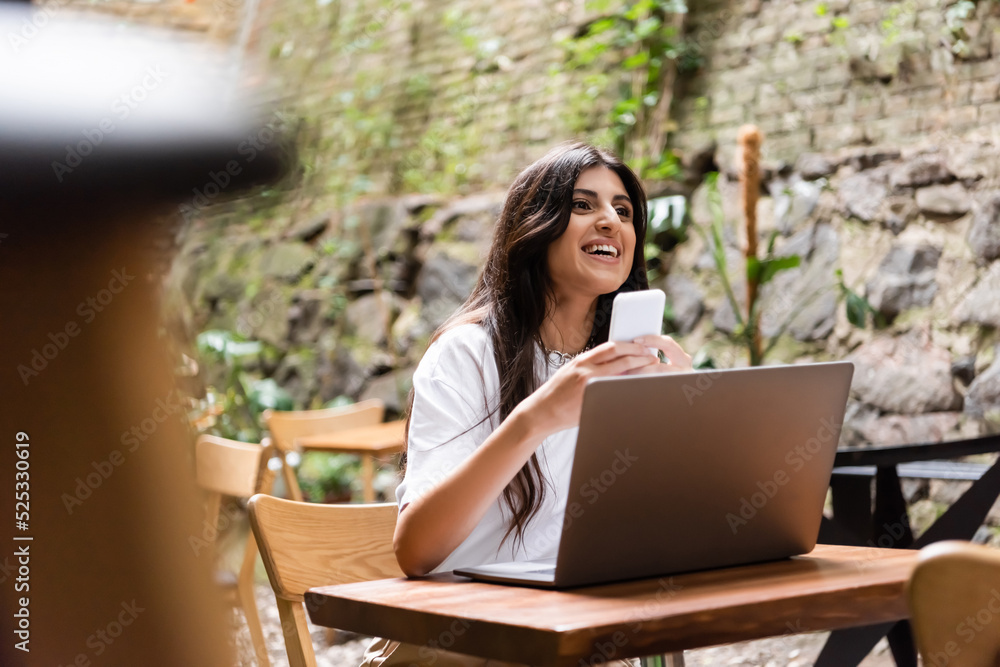 Smiling woman holding smartphone near laptop and looking away in outdoor cafe Smiling woman holding smartphone near laptop and looking away in outdoor cafe