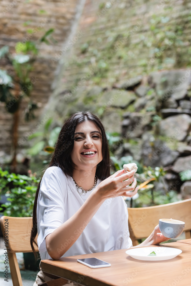 Happy woman holding coffee and dessert and smiling at camera near smartphone on terrace of cafe Happy woman holding coffee and dessert and smiling at camera near smartphone on terrace of cafe
