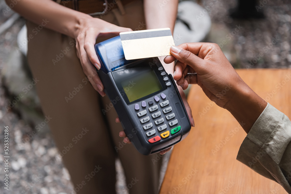 Cropped view of waitress holding payment terminal near african american client with credit card on cafe terrace Cropped view of waitress holding payment terminal near african american client with credit card on cafe terrace