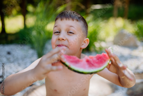 Little boy sitting near lake and eating watermelon on hot sunny day during summer vacation.
