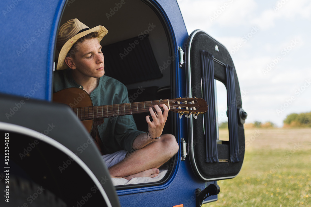 Naklejka premium Young man sitting in a van, camping ang playing at guitair.