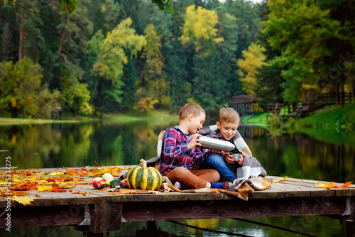 A boy pours tea from a thermos by the lake on the pier. Friends on a picnic in nature in autumn. The children wrapped themselves in a blanket.