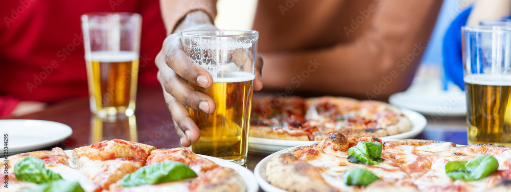 Horizontal banner or header with close-up of african guy's hand taking glass of beer at pizza restaurant - Multi ethnic Friends having fun eating pizza and drinking beer at pub