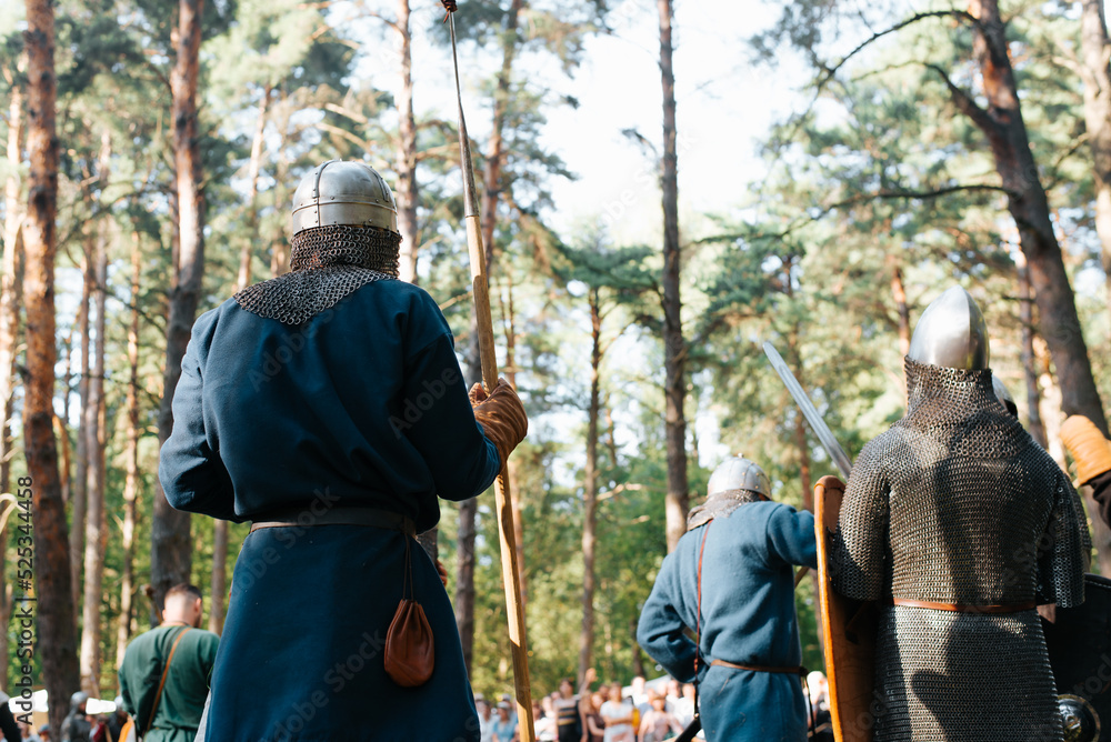 Vikings and knights in helmets and chain mail holding swords and spears standing outdoors, back view. Costumed festival of medieval culture, historical reenactment. Vikings and knights in helmets and chain mail holding swords and spears standing outdoors, back view. Costumed festival of medieval culture, historical reenactment.