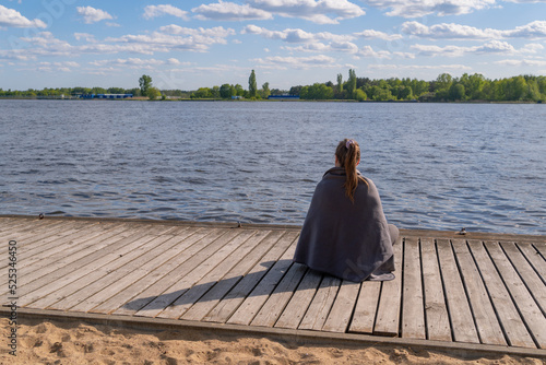 a woman sits on the edge of a pier made of wooden bars, a woman covered with a warm gray blanket looks into the distance at the blue water of the lake on the other shore, a forest strip, sunny, windy  © Tania