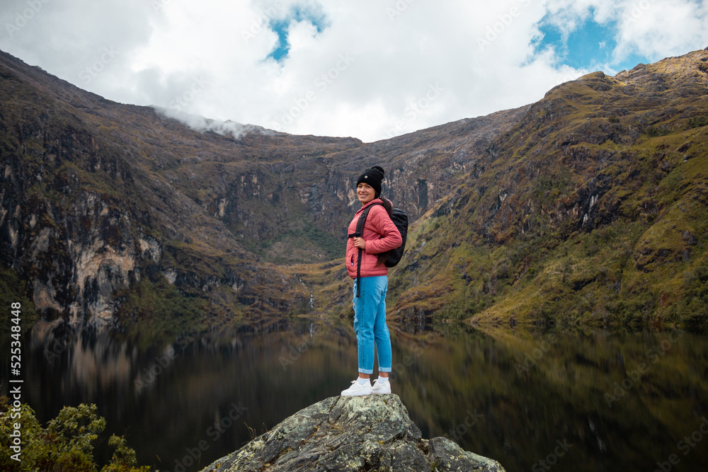 Una chica con una chaqueta rosa y mochila parada  junto a un tranquilo lago en las hermosas montañas - obrazy, fototapety, plakaty 
