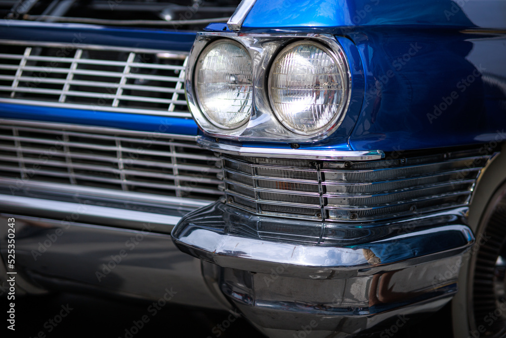 The front quarter panel, headlight housings, and chrome grill and bumper, and white wall tire of a vintage classic blue auto.