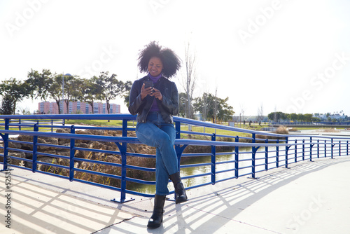 Young, beautiful Afro-American woman with mobile phone shopping online, checking her email and social networks in the park. The woman is wearing casual and comfortable clothes and is happy.
