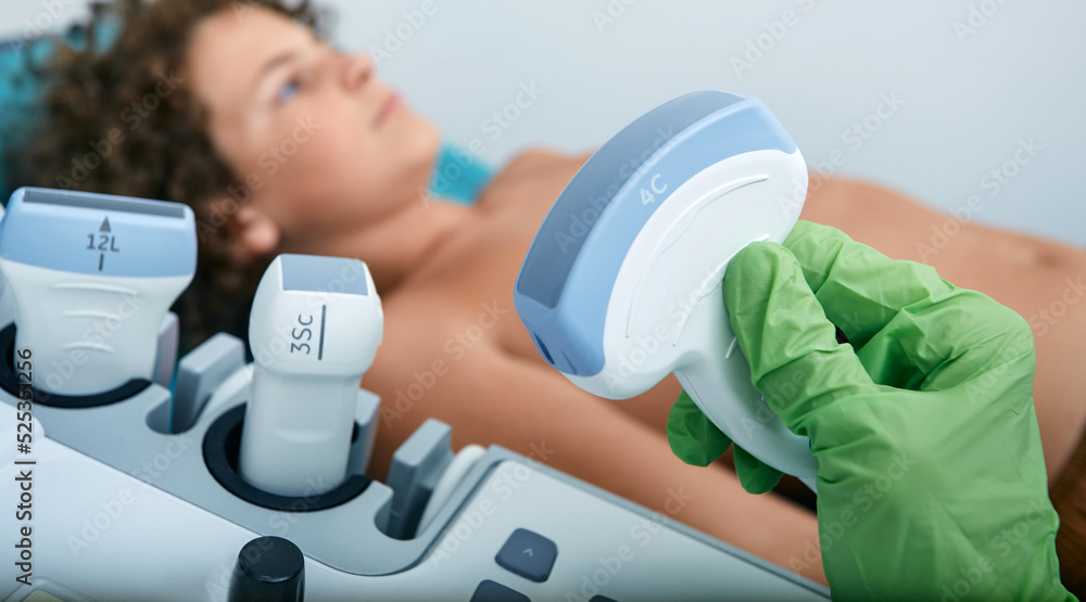 Ultrasound exam for child. Ultrasound probe in doctor's hands before sonography procedure for curly boy lying on bed in medical clinic Ultrasound exam for child. Ultrasound probe in doctor's hands before sonography procedure for curly boy lying on bed in medical clinic