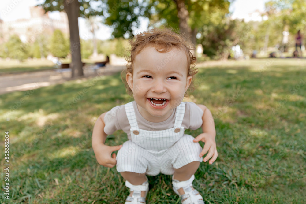 Outdoor portrait of lovely smiling cute little girl with curls having fun and posing at camera on background of green lawn. Happy kid spend time outdoor in warm sunny day 