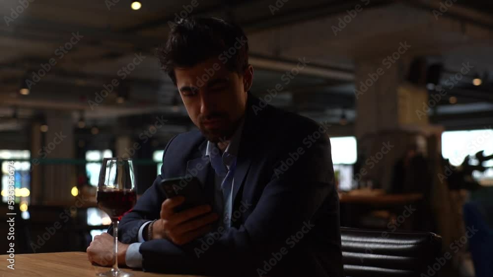 Close-up portrait of cheerful young man in suit using smartphone chatting with friends and drinking red wine sitting at table in restaurant. Handsome bearded male waiting for girlfriend on date. Close-up portrait of cheerful young man in suit using smartphone chatting with friends and drinking red wine sitting at table in restaurant. Handsome bearded male waiting for girlfriend on date.