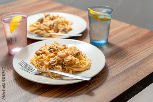 Two plates on the edge of a wooden table. Pasta with mussels and shrimp, water with lemon.