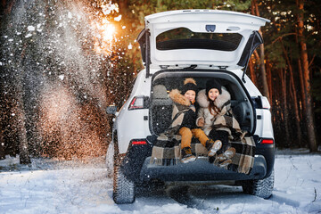 Two happy children on walk in winter forest. Children sit in trunk of family car. Children warm themselves wrapped in a warm blanket.