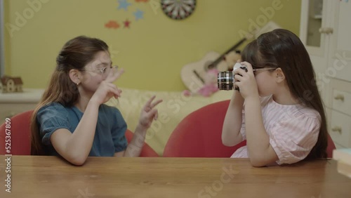 Little beautiful and cute girls are sitting at the table in the nursery and taking pictures of each other. Loving girls are chatting and laughing while sitting in their armchairs in their happy home.