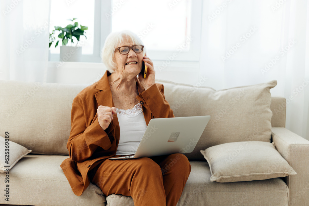 a sad, angry elderly woman in a brown suit is sitting with a laptop on her lap on a cozy sofa working from home and talking on the phone, looking serious and holding her hand in her fist a sad, angry elderly woman in a brown suit is sitting with a laptop on her lap on a cozy sofa working from home and talking on the phone, looking serious and holding her hand in her fist