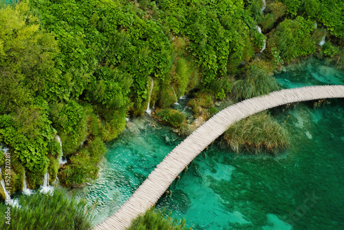 Hiking tourists walk on wooden footbridge surrounding lake with turquoise water in natural reserve. Majestic view of blue lake among picturesque landscape on Plitvice lakes upper view