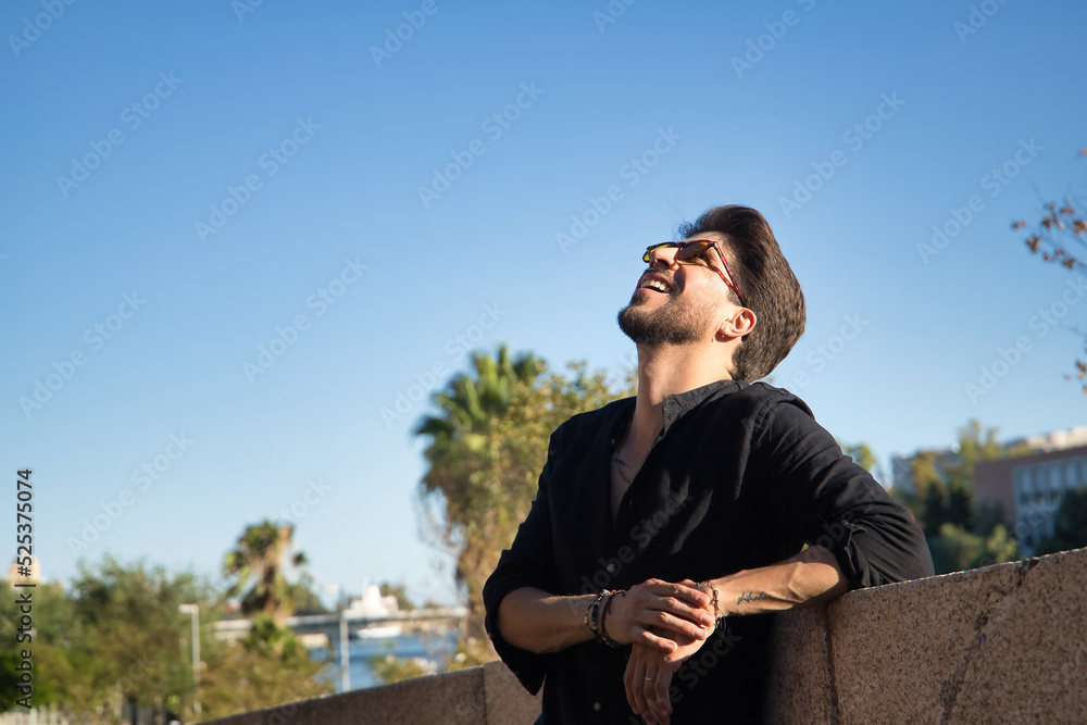 Young Hispanic man with a beard, sunglasses and black shirt, looking up at the sky smiling happily, leaning on a stone wall. Model concept, beauty, fashion, triumph, happiness.