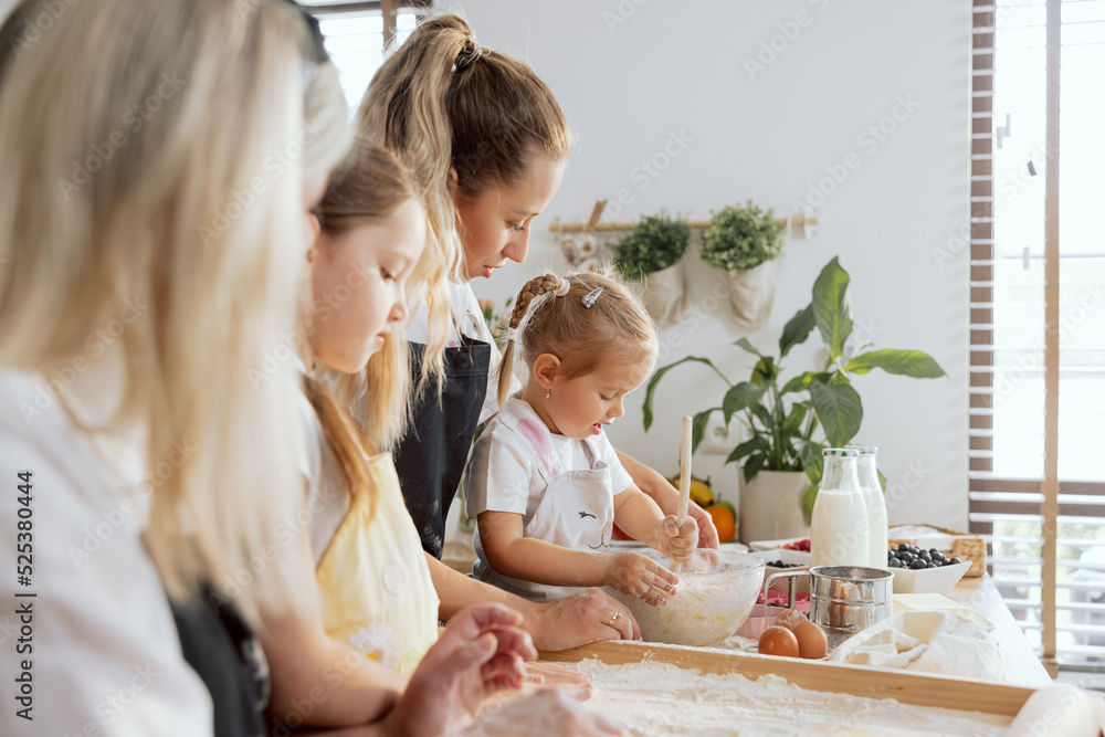 Curious little daughter kneading dough with spoon in large bowl young mother managing process. In foreground silouhette of elderle granny and older sister watching process.