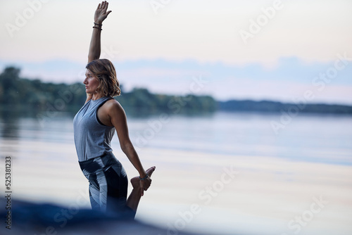 Valokuvatapetti A woman is practicing yoga and balancing on one foot on the dock near the river in nature