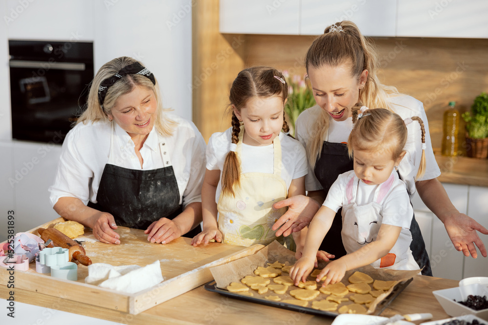 Young mother granny and curious daughters helping baking cookies using cookie cutters. Preschooler daughter placing cookies biscuits on the parchment girls having fun together watching the process Young mother granny and curious daughters helping baking cookies using cookie cutters. Preschooler daughter placing cookies biscuits on the parchment girls having fun together watching the process