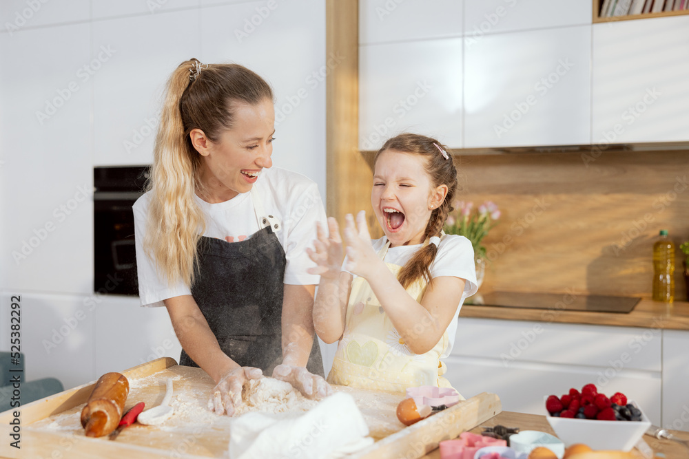 Beautiful older sister with younger one having fun in moder light kitchen cooking baking cake cookies biscuits. Kneading dough. Younger girl clapping in hands with flour.