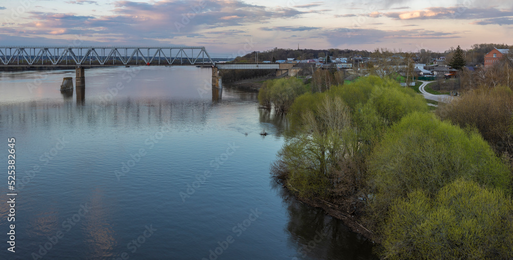 Evening landscape, sunset on the river. Spring, green trees in the foreground. View of the industrial area of the railway bridge. Outskirts.
