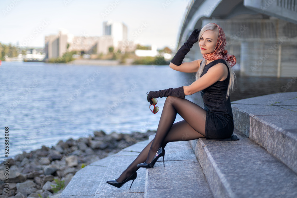 Beautiful blonde girl in black dress with perfect legs in pantyhose and  shoes with high heels posing outdoor near the river bridge in the rays of  sun. Street fashion photo. Photos