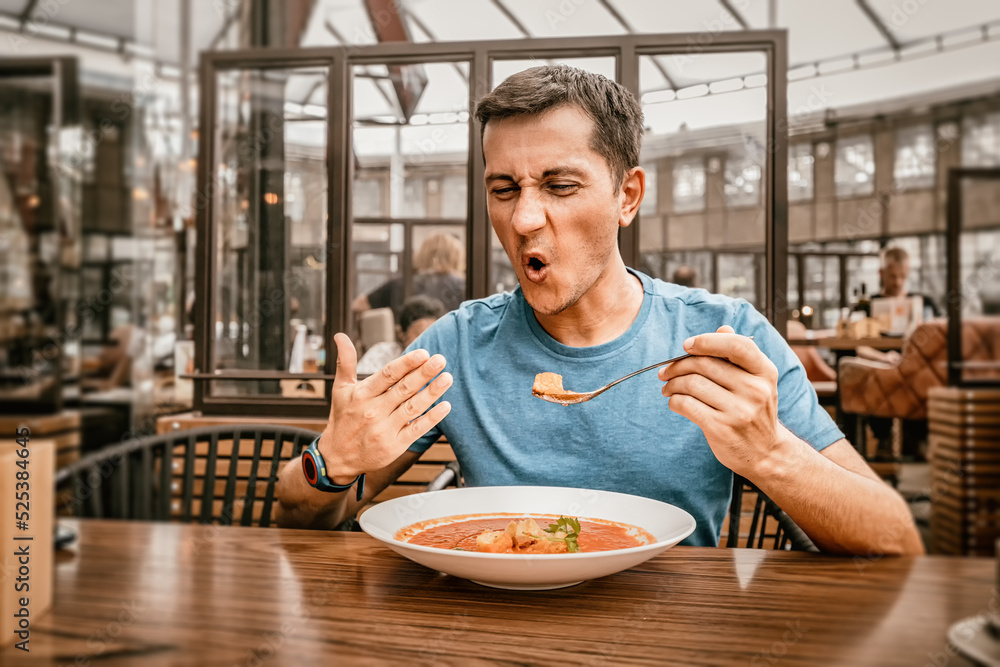 A man tries a spicy and hot red soup in a restaurant and reacts funny emotionally. Seasonings in the national cuisine and an unhealthy diet with overabundance of pepper