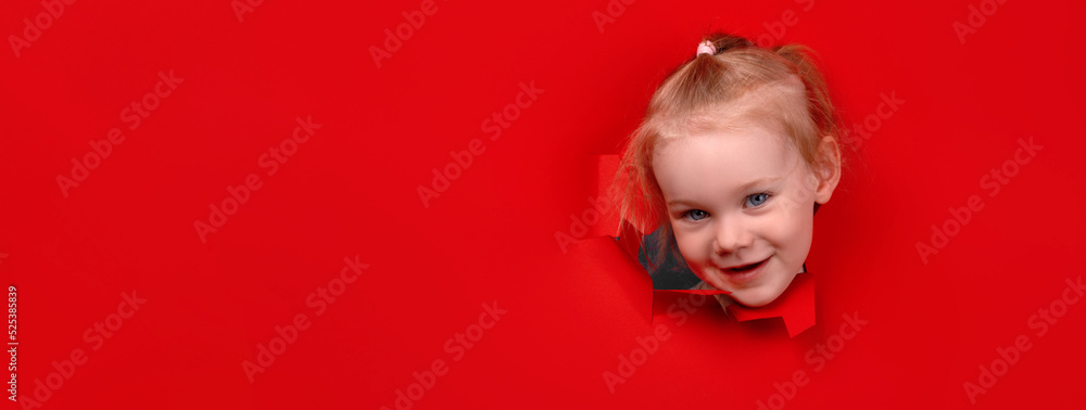 Portrait of young smiling girl looking through hole in the red paper wall, panoramic layout Portrait of young smiling girl looking through hole in the red paper wall, panoramic layout