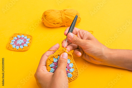 Closeup of woman's hands with a hook inserted in crochet element with the backgound of yellow table, yellow skein of yarn and finished granny square element photo
