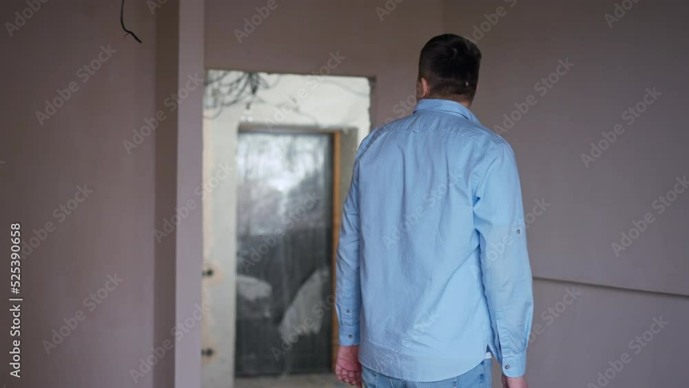Thoughtful young man looking around standing in apartment under construction gesturing thumb up smiling looking at camera. Portrait of satisfied owner posing in new flat Thoughtful young man looking around standing in apartment under construction gesturing thumb up smiling looking at camera. Portrait of satisfied owner posing in new flat