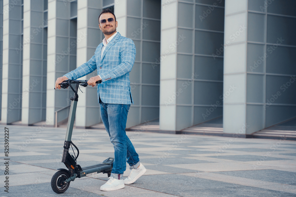 Joyful businessman with scooter dressed in blue suit and sunglasses against city building. Joyful businessman with scooter dressed in blue suit and sunglasses against city building.