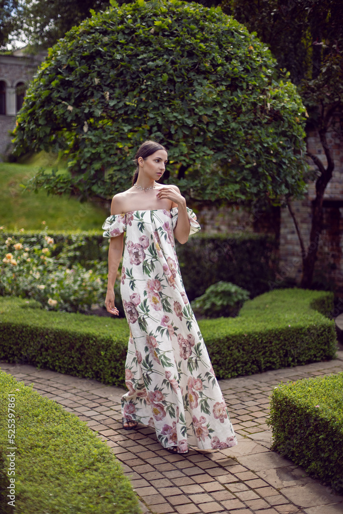 Outdoor portrait of a beautiful luxury brunette woman in a dress with flowers stands in a park with trimmed trees.