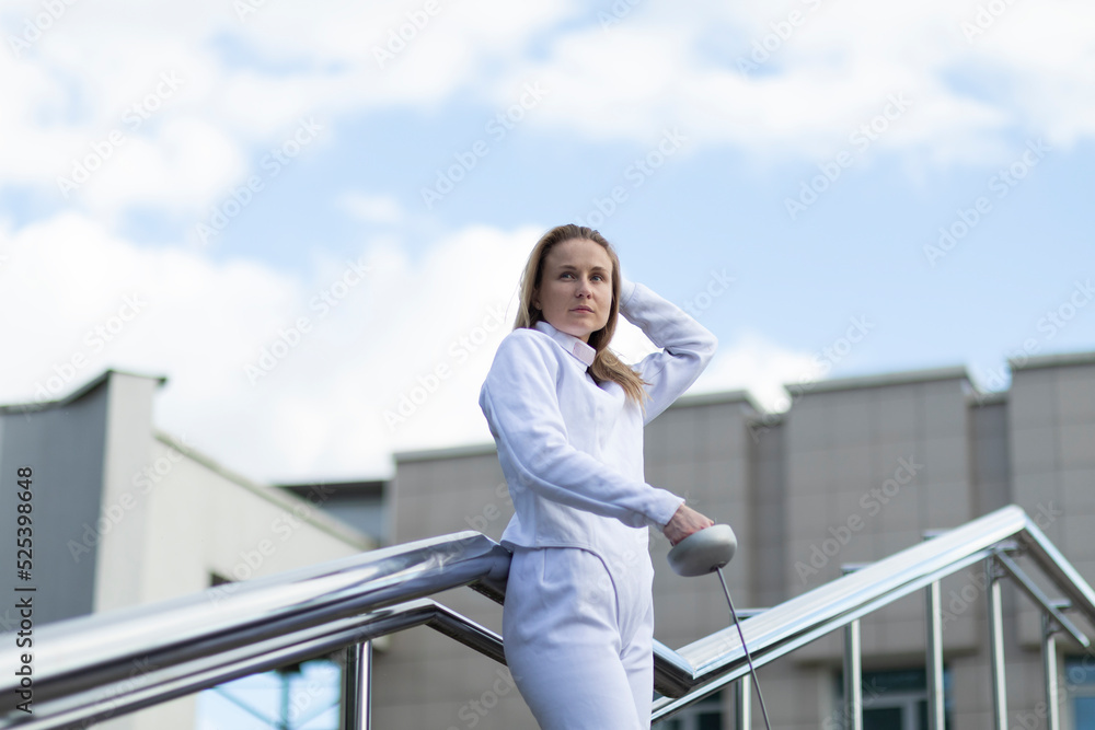 Young beautiful woman of Millennial Generation wears white fencing costume, holds metal pee in hand outside, stays on stairs, sky is on background. Horizontal plane. Sport, hobby concept.