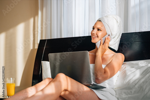 Pretty woman wrapped in bath towel lying on bed with laptop on knees and holding smartphone in hands. Lifestyles with modern technology. A woman works on vacation at the hotel  after a bath or shower.